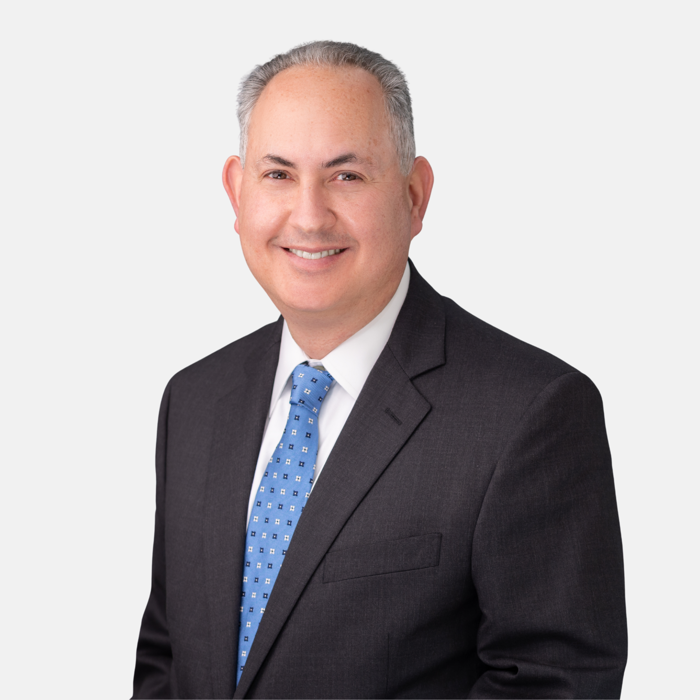 Headshot of Gary P. Seligman with short grey hair, wearing a dark suit jacket, white shirt, and blue pattern tie, smiling against a light background