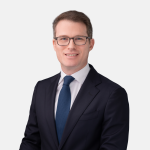 Headshot of John E. Howell with short brown hair and glasses, wearing a dark suit, light shirt, and blue tie, smiling against a light background
