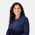 Headshot of Kimberly M. Melvin with long dark curly hair, wearing a dark blue blazer, patterned top, and gold chain necklace, smiling against a light background